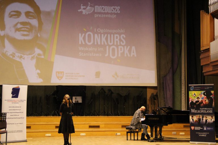 Na zdjęciu kobieta na scenie śpiewa. Z prawej strony mężczyzna gra na fortepianie.