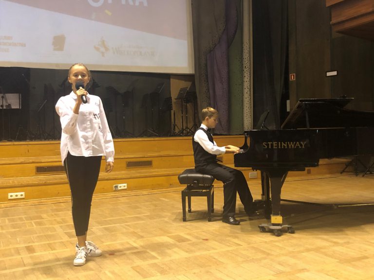 Na zdjęciu młoda dziewczyna na scenie śpiewa. Obok chłopak gra na fortepianie.