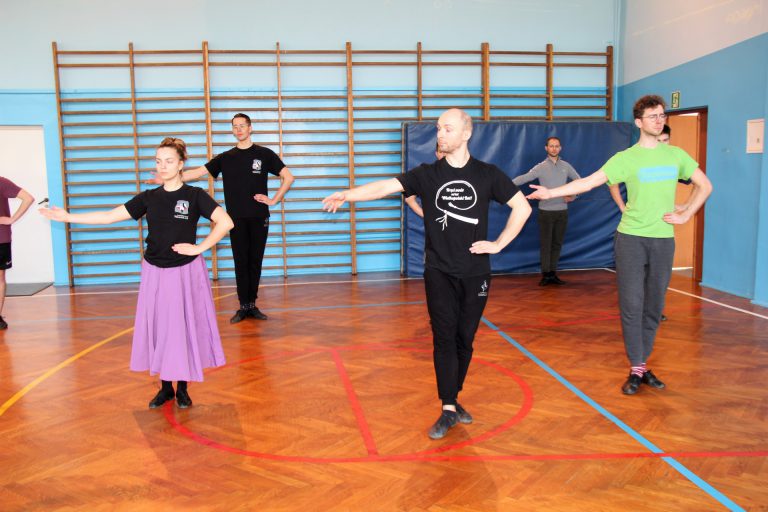 Tancerze w strojach ćwiczebnych wykonują ćwiczenia rąk w pozycji stojącej.
