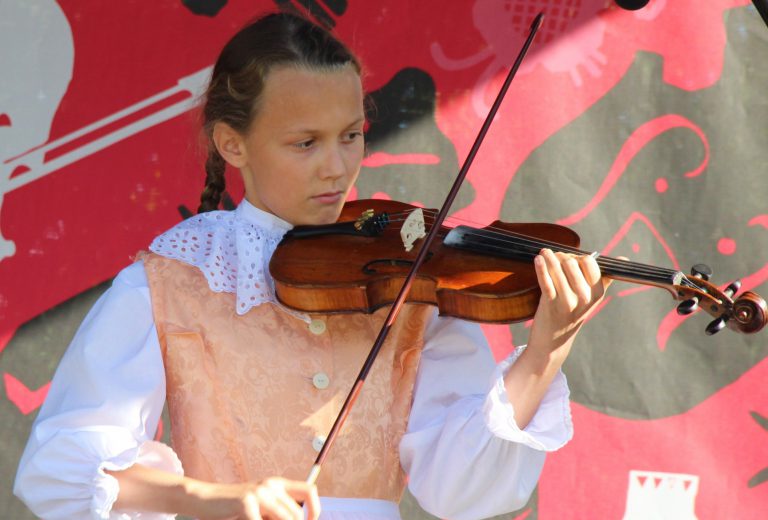 Na zdjęciu młoda dziewczyna w stroju ludowym gra na skrzypcach.