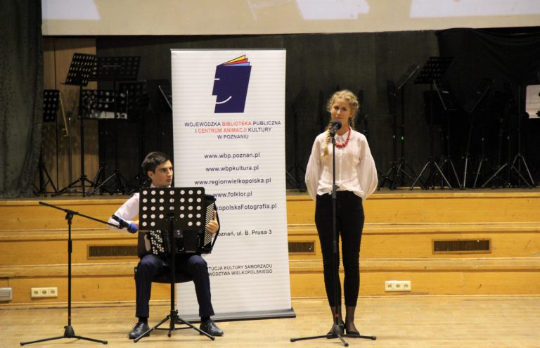 Na zdjęciu młoda dziewczyna śpiewa na scenie. Za nią mężczyzna gra na akordeonie.