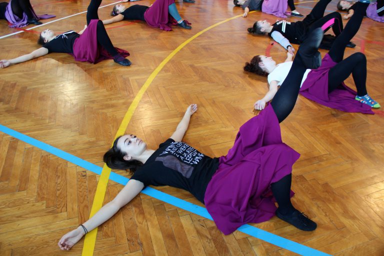 Tancerze w strojach ćwiczebnych leżą na plecach i wykonują ćwiczenia nóg.