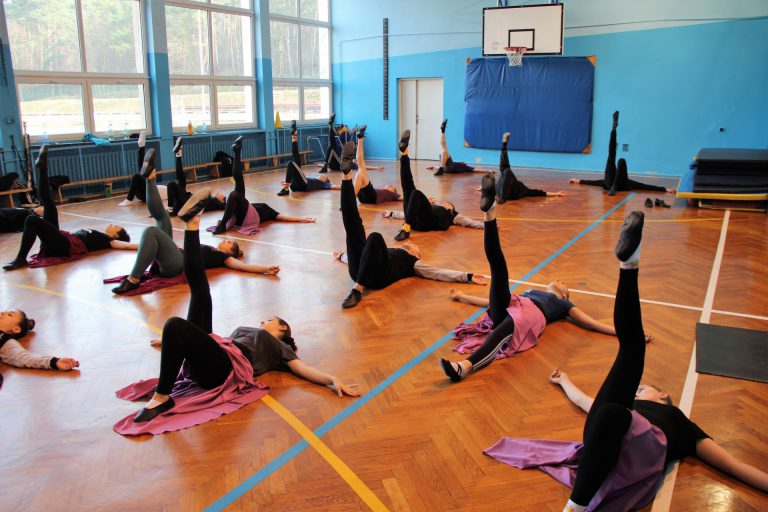 Tancerze w strojach ćwiczebnych leżą na plecach i wykonują ćwiczenia nóg.