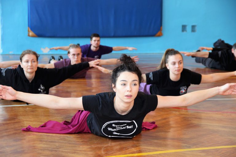 Tancerze w strojach ćwiczebnych wykonują ćwiczenia pleców w pozycji leżenia na brzuchu.