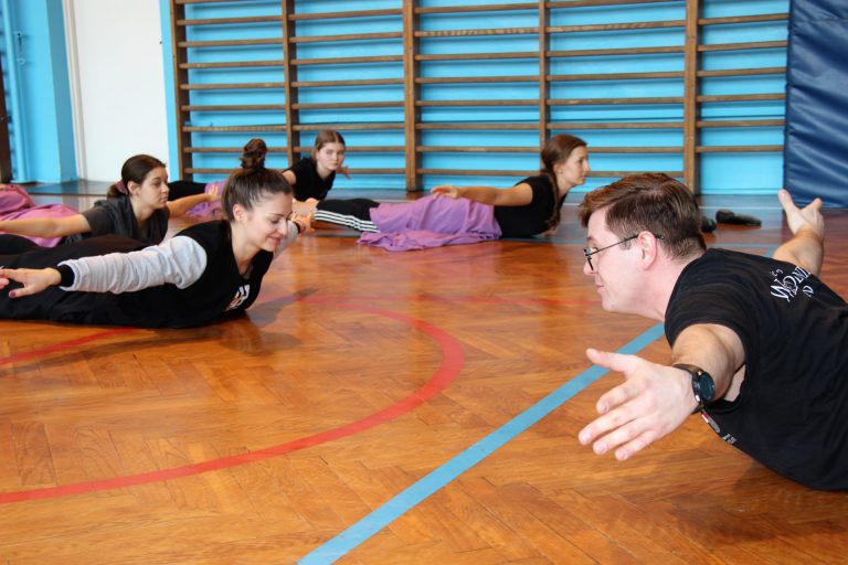 Tancerze w strojach ćwiczebnych wykonują ćwiczenia pleców w pozycji leżenia na brzuchu.