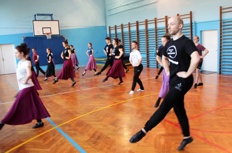 Tancerze w strojach ćwiczebnych wykonują ćwiczenia nóg w pozycji stojącej.