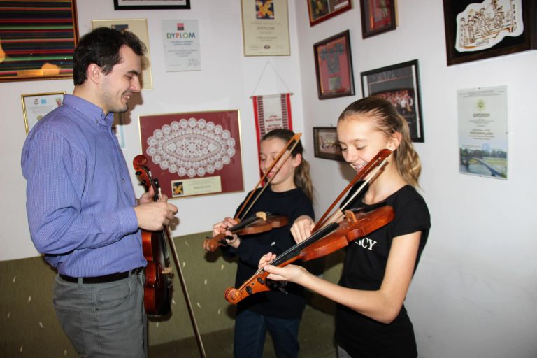 Na zdjęciu mężczyzna ze skrzypcami w ręce stoi naprzeciwko dwóch dziewczynek i się uśmiecha. Dwie dziewczynki na zdjęciu grają na mazankach.